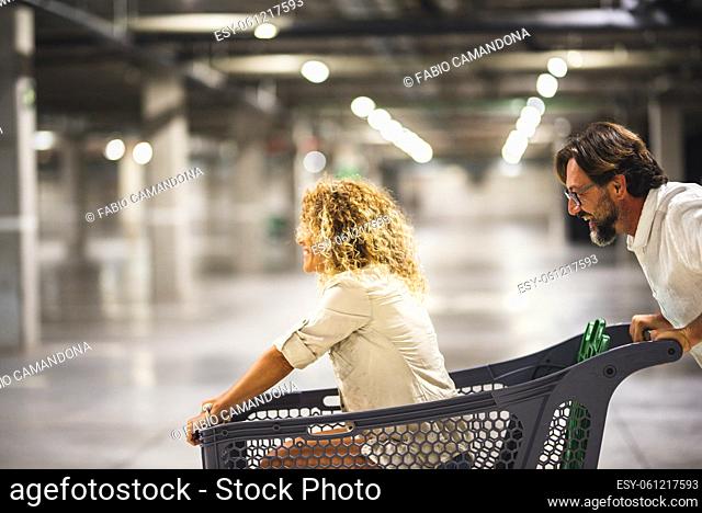 Happy couple having fun with shopping cart at parking garage, Husband pushing his carefree wife inside shopping trolley. Enthusiastic couple with shopping cart...