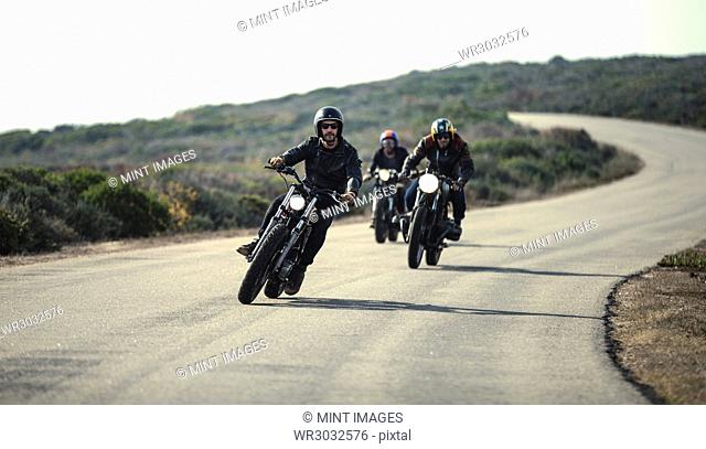 Three men wearing open face crash helmets and sunglasses riding cafe racer motorcycles along rural road