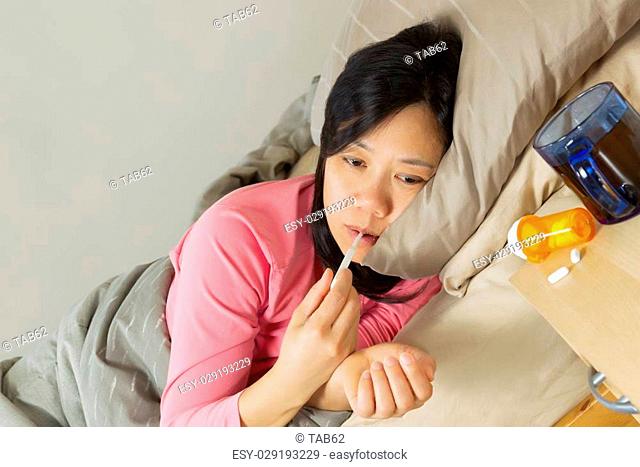 Horizontal photo of mature woman holding thermometer in mouth while lying in bed sick