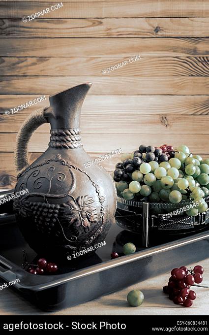 On the table, in a glass vase with green grapes next to a ceramic jug of wine. Presented on a wooden background. Copy space