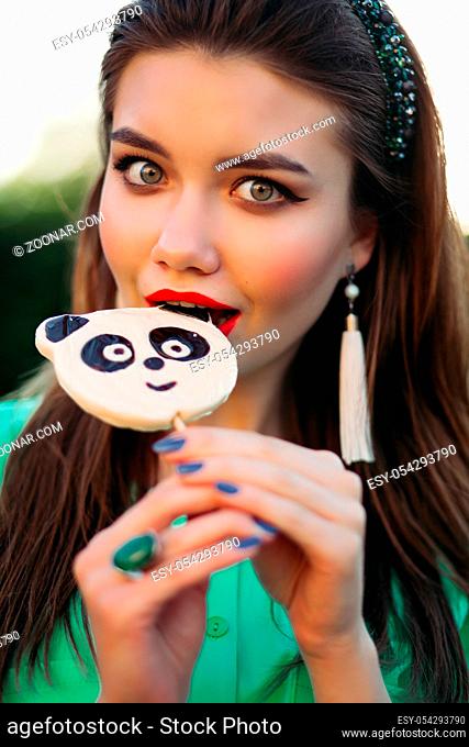 Portrait of beautiful brunette girl in green dress, eating candy like panda on stick and looking at side. Pretty and stylish woman with red lips