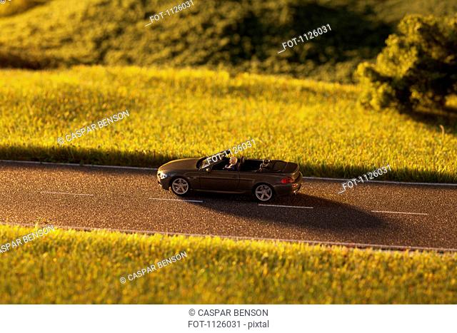 A diorama of a miniature man driving a toy convertible sports car on a rural road