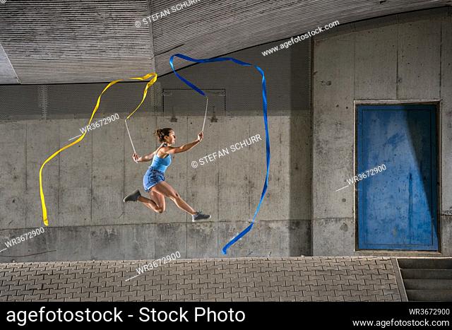 Young woman holding ribbons practicing rhythmic gymnastics against concrete wall under bridge