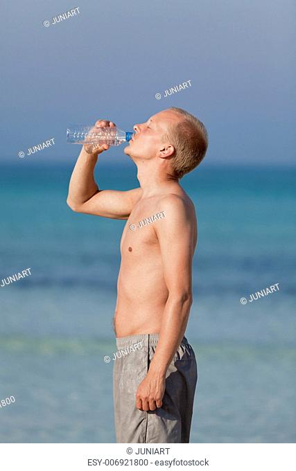 Young man boy drinking refreshing water from a bottle on the beach by the sea in summer vacation