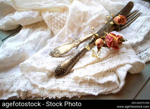 Light pastel colored tableware set: vintage silverware on napkin and delicate pink dry roses on rustic shabby wooden table