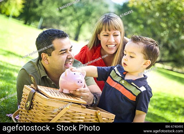 Happy mixed-race couple give their son a piggy bank at a picnic in the park