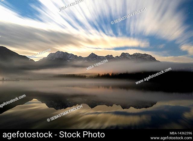 Nighttime long exposure of the clouds and fog in autumn, with the Karwendel mountains reflected in the clear water of the Barmsee near Krün