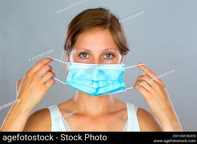 Young woman putting on a surgical mask in studio. Female holding a blue respiratory in front of face. Blonde girl looking to the camera and covering mouth and...