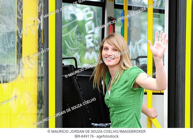 Young, pretty, blonde woman wearing jeans and a T-shirt getting on a yellow tram and waving good bye