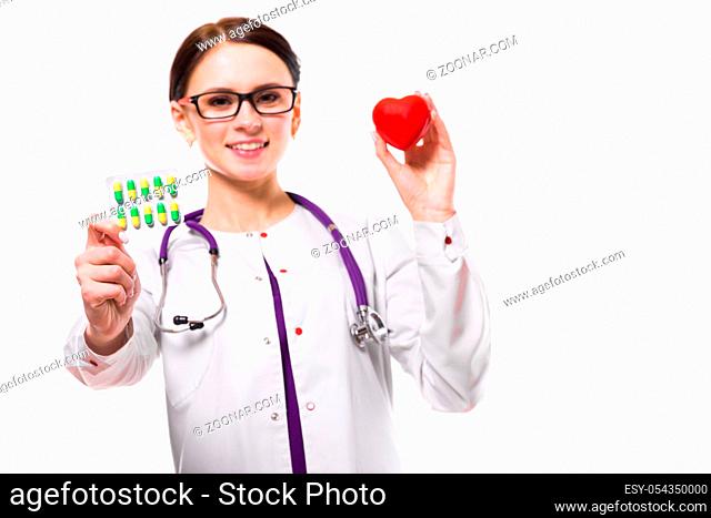 Portrait of young attractive positive friendly smiling woman doctor standing in office with heart and pills in her hands looking in camera in white uniform...