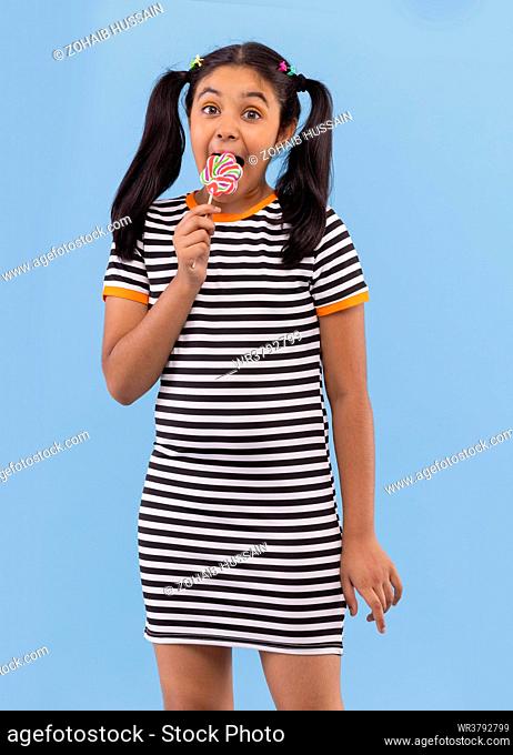Portrait of little girl licking lollipop against plain background