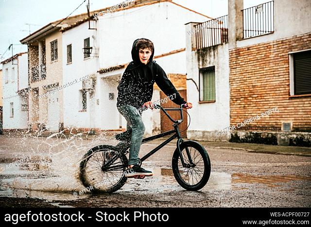 Teenage boy splashing water in puddle while cycling on street during rainy season