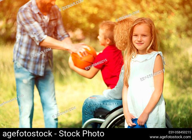 Beautiful Blonde Girl On Foreground Is Looking At Camera And Smiles. Blurred Image Of Man Throwing Ball To Little Boy, Who Sits On The Knees Of His Disabled...