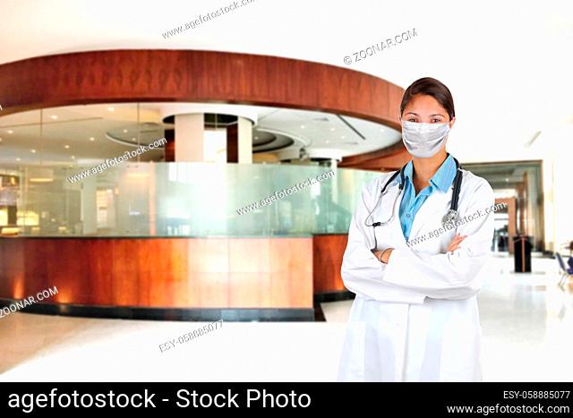 Young woman nurse or doctor wearing surgical mask and Lab Coat with arms crossed and Stethoscope draped around her neck, in medical facility lobby