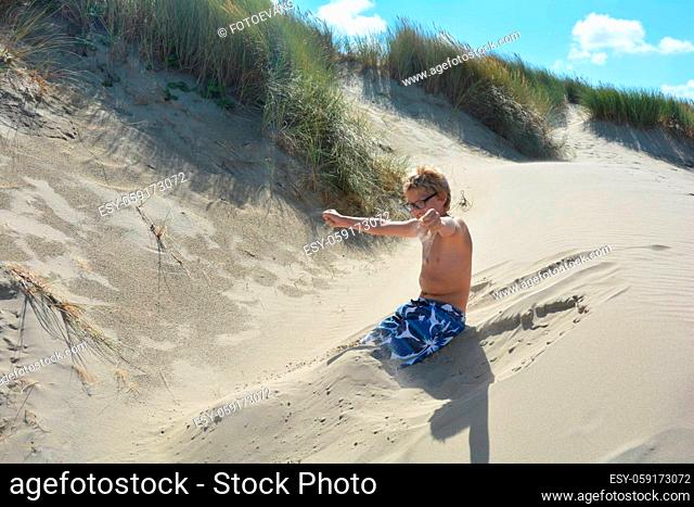 Boy sits in the sand in the dunes with swimming trunks and lets the sand trickle out of his hands, Legs dug in
