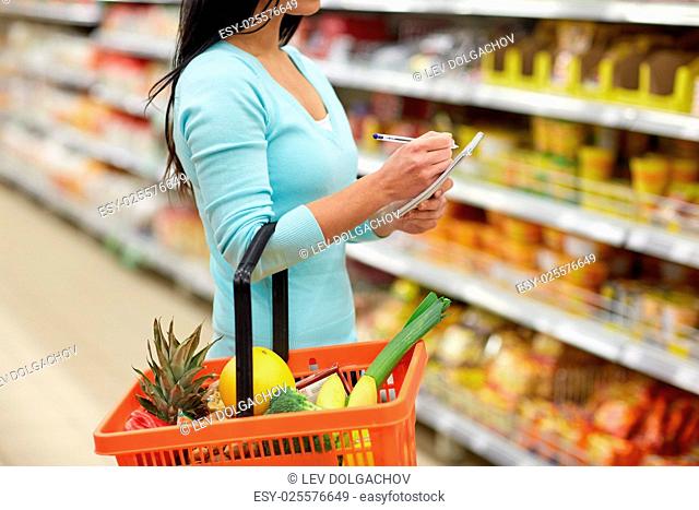 consumerism and people concept - woman with notebook and shopping basket buying food at grocery store or supermarket