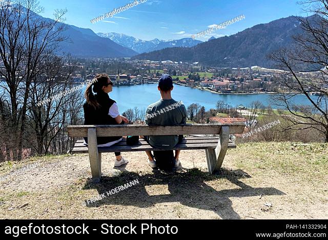 Young couple takes a rest on a bench. Hikers on the Hoehenweg over the Tegernsee with a view of Rottach Egern on April 1st, 2021