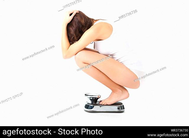 Diet and weight, young woman sitting on her haunches on a scale, she is desperate