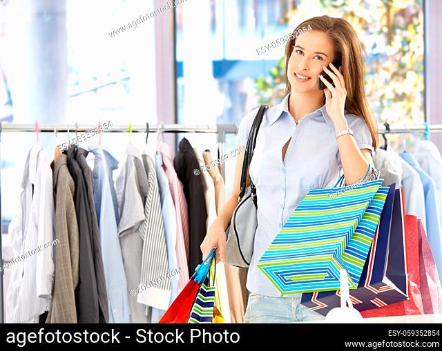 Woman on mobile phone call standing in clothes shop, holding shopping bags, smiling