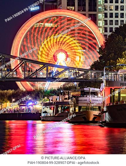 03 August 2019, Hessen, Frankfurt/Main: A Ferris wheel turns on the banks of the Main, while the lights of the Mainfest are reflected in the river in front of...