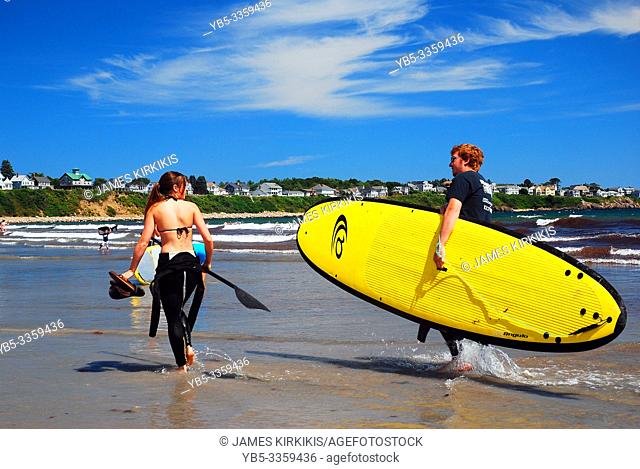 A young man carries the surfboard for his better half as she prepares to catch a wave