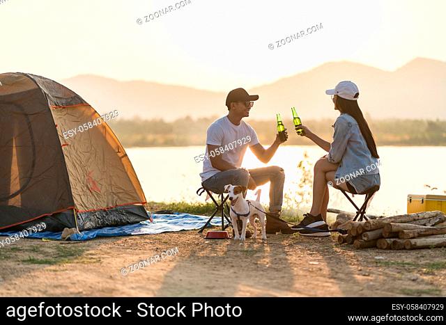 Young adult Asian couple drinking beer beside their tent campsite while sunset. Camping trip with dog outdoor activity campsite concept