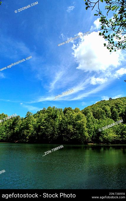 View of smooth lake surrounded by forest among mountains, on cloudy sky background