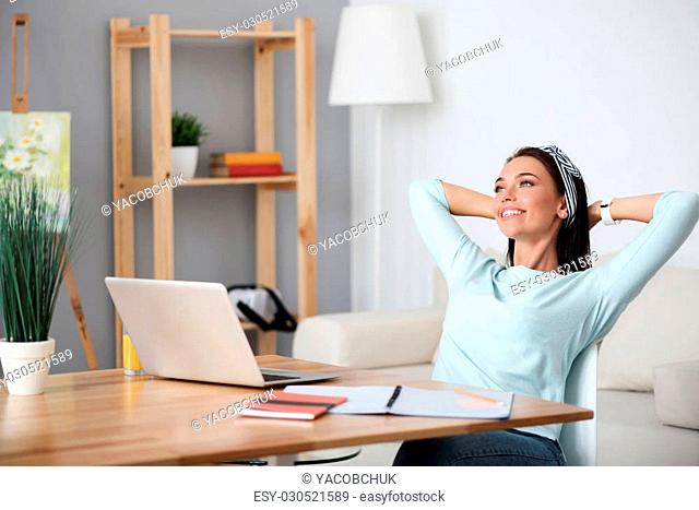 Get some rest. Cheerful relaxed woman smiling and sitting at the table while holding her hands behind the head