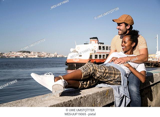 Happy young couple relaxing on a wall at the waterfront, Lisbon, Portugal