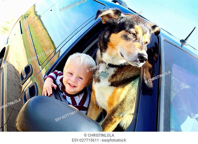 a cute, smiling baby boy and his German Shepherd dog are hanging out a minivan window on a summer day
