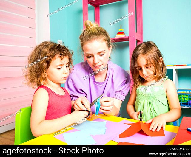 The woman teacher with two little girls cuts out colored paper. The tutor with girls are sitting at a table in the kindergarten