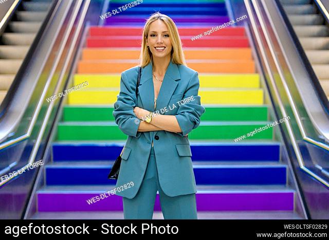 Smiling blond working woman standing with arms crossed in front of colorful steps