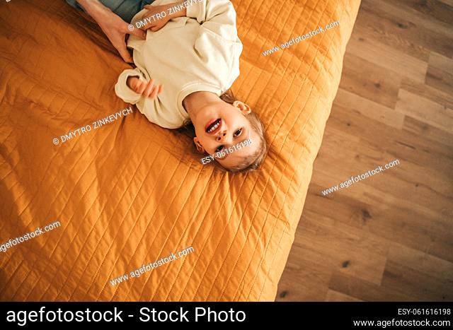 Top view of a calm cute little girl lying on the quilted bedspread in the bedroom and looking up