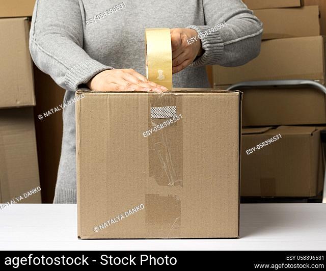 woman in a gray sweater holds a roll of duct tape and packs brown cardboard boxes on a white table, behind a stack of boxes. Moving concept