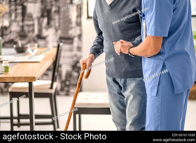 Cropped photo of a nurse in uniform supporting an old man with a walking stick