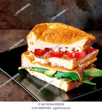 Low-carb gluten free Cloud bread veggie sandwich with spinach, avocado, feta cheese, tomatoes and pesto sauce, served on black plate over dark wooden textured...