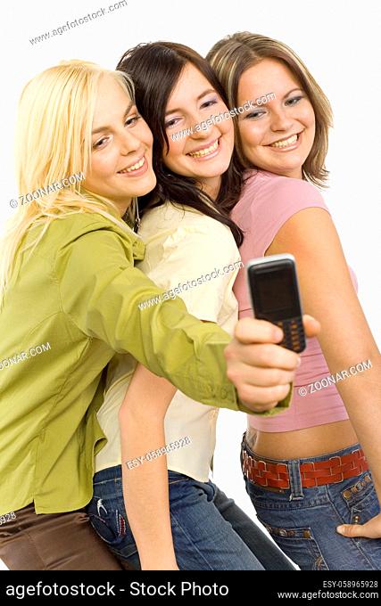 Three girl friends are standing. One of them is holding mobile phone and making the picture. Isolated on white in studio