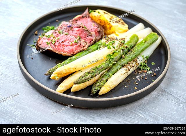 Traditional white and green asparagus with barbecue dry aged sliced beef fillet and fried potatoes served as closeup on a modern design plate