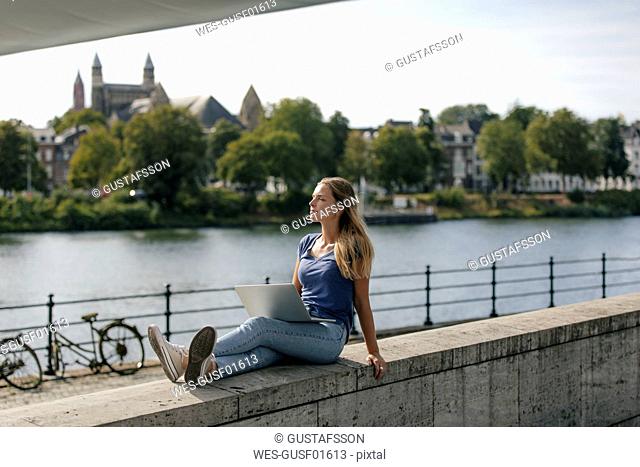 Netherlands, Maastricht, young woman sitting on a wall at the riverside with laptop