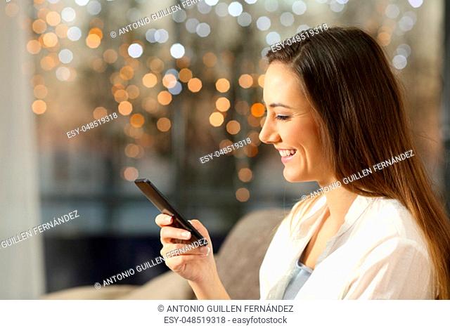 Side view portrait of a happy woman using a smart phone sitting on a sofa at home in the evening with little lights in the background