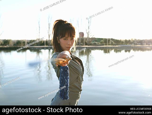 Confident female martial artist holding sword while standing against lake on sunny day