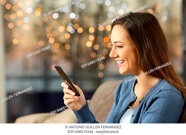 Side view portrait of a happy girl using a smart phone sitting on a sofa in the living room at home with lights in the background