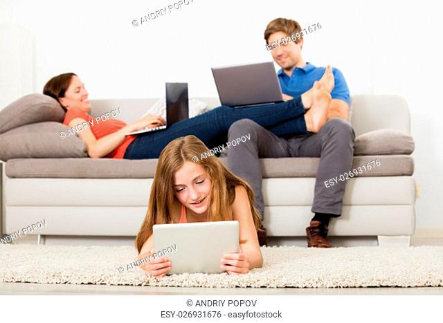Girl Lying On Carpet Using Digital Tablet In Front Of Her Parent Working On Laptop