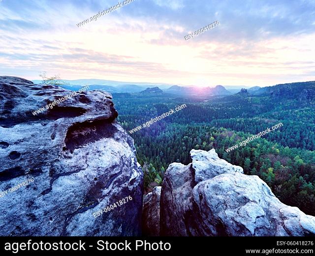Spring morning nature. The cracked sandstone cliff above forest valley, daybreak Sun at horizon. Hills increased from misty background