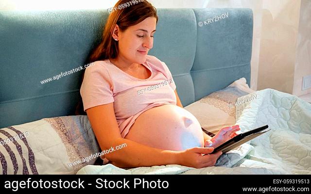 Smiling pregnant woman in pajamas using tablet computer and typing message while lying in bed at night