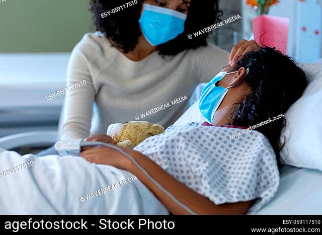 Mixed race mother and sick daughter in face masks in hospital, girl sleeping and holding teddy bear