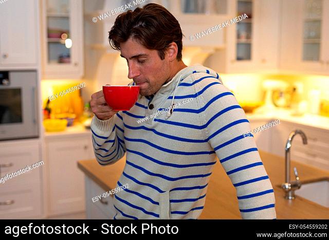 Side view of a Caucasian man at home wearing a blue striped sweater, standing in the kitchen, drinking a cup of coffee