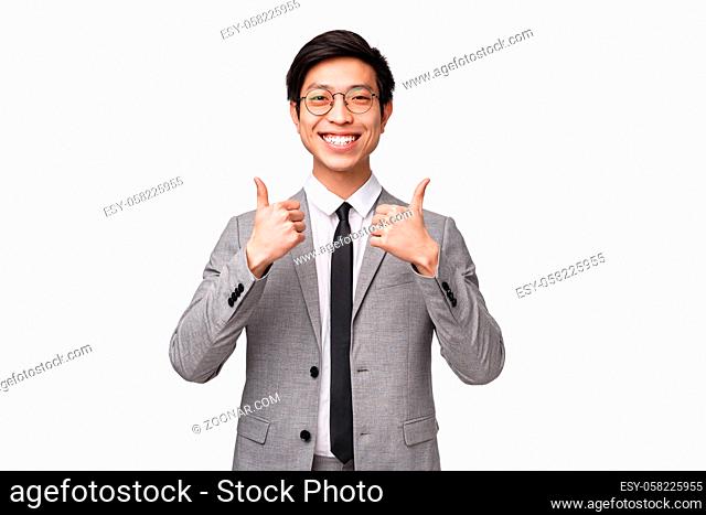 Waist-up portrait of pleased, fully satisfied asian young man in grey suit, showing thumbs-up and smiling with approval, like awesome concept