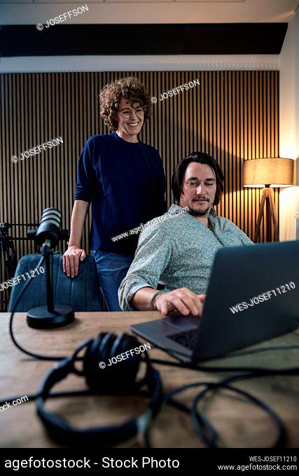 Smiling presenter talking with colleague using laptop in recording studio
