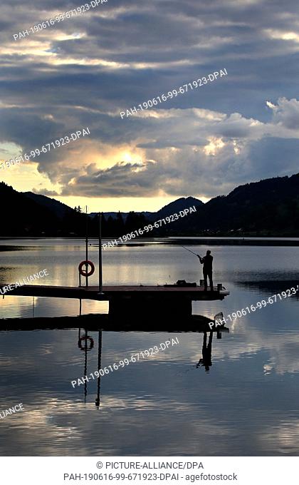 16 June 2019, Bavaria, Immenstadt: An angler on a jetty is reflected in the water of the Großer Alpsee shortly before sunset